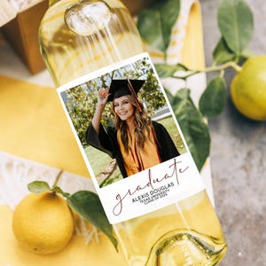 May include: A clear wine bottle with a custom label featuring a graduation photo. The label reads "graduate" in cursive, along with the graduate's name and university. The bottle is partially filled with white wine.