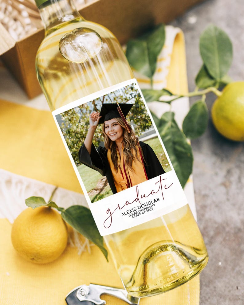 May include: A clear wine bottle with a custom label featuring a graduation photo. The label reads "graduate" in cursive, along with the graduate's name and university. The bottle is partially filled with white wine.