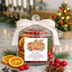 May include: A clear glass jar filled with cranberries, orange slices, and cinnamon sticks, topped with a white ribbon. A square label features a holiday illustration and text: Christmas STOVE TOP POTPOURRI. ADD ALL INGREDIENTS TO A SAUCE PAN WITH ENOUGH WATER TO COVER, SIMMER ON LOW HEAT. ADD WATER AS NEEDED. ENJOY! THE HILLARD FAMILY.