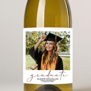May include: A clear wine bottle with a white label featuring a graduation photo. The label includes the word "graduate" in a cursive font, along with the graduate's name, university, and class year. The graduate is wearing a cap and gown.