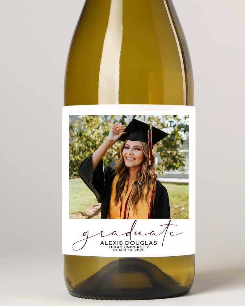 May include: A clear wine bottle with a white label featuring a graduation photo. The label includes the word "graduate" in a cursive font, along with the graduate's name, university, and class year. The graduate is wearing a cap and gown.