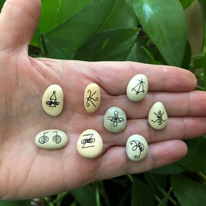 May include: Eight smooth, oval-shaped stones with different black ink symbols drawn on them. The stones are in a hand, palm up.
