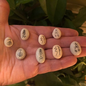 以下が含まれることがあります： それぞれ異なる黒いシンボルまたは文字が刻まれた、滑らかな楕円形の石が8つ。オフホワイトの石は手で持たれており、背景には緑の葉があります。シンボルは日本の起源のようです。