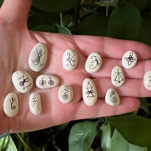May include: A selection of smooth, cream-coloured stones, each decorated with distinct black ink symbols. The stones are of various shapes and sizes, with the symbols appearing hand-drawn. The arrangement suggests a spiritual or decorative purpose.