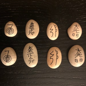 May include: Eight smooth, oval stones with black symbols and characters. The stones are beige and arranged on a dark wooden surface. The symbols include a spiral and various Japanese characters.