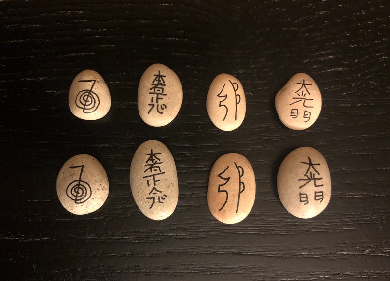 May include: Eight smooth, oval stones with black symbols and characters. The stones are beige and arranged on a dark wooden surface. The symbols include a spiral and various Japanese characters.