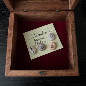 May include: Open wooden box with a red velvet interior. Inside, a yellow sticky note reads "Intentions Wishes Photos." Four small stones with Japanese characters are arranged on the note, suggesting a theme of mindfulness or meditation.