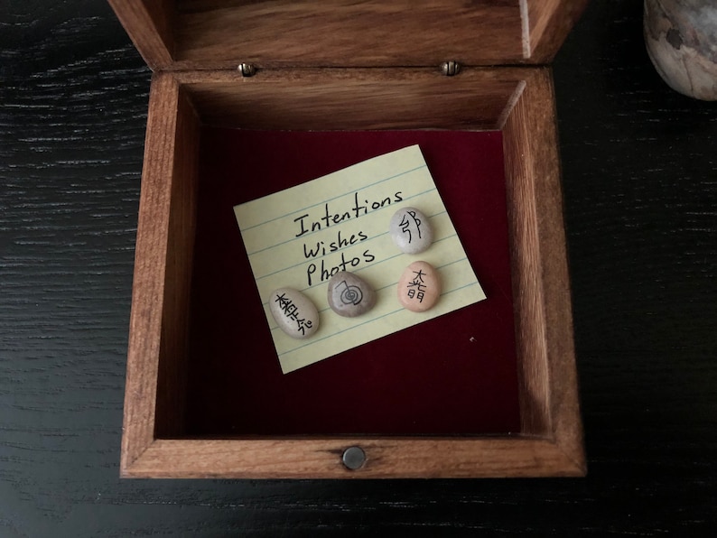 May include: Open wooden box with a red velvet interior. Inside, a yellow sticky note reads "Intentions Wishes Photos." Four small stones with Japanese characters are arranged on the note, suggesting a theme of mindfulness or meditation.