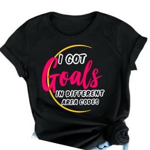Könnte beinhalten: Schwarzes T-Shirt mit dem Text "I GOT Goals IN DIFFERENT AREA CODES". Das Wort "Goals" ist in rosa Schrift mit gelbem Halbmond-Umriss geschrieben. Der restliche Text ist weiß.