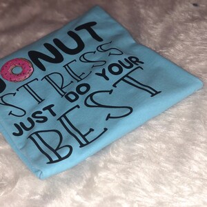 May include: Light blue t-shirt with the text "Donut stress just do your best" in black lettering. A pink and white donut graphic is above the text.