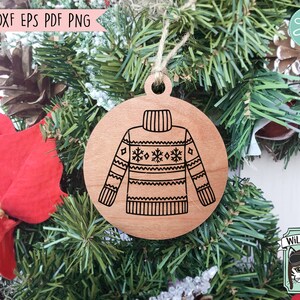 Op de afbeelding: Een houten kerstversiering in de vorm van een cirkel met een zwart-witte illustratie van een trui met een sneeuwvlokkenpatroon. De versiering hangt aan een groene kerstboom.