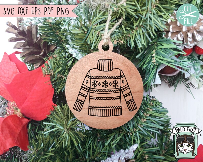 Op de afbeelding: Een houten kerstversiering in de vorm van een cirkel met een zwart-witte illustratie van een trui met een sneeuwvlokkenpatroon. De versiering hangt aan een groene kerstboom.
