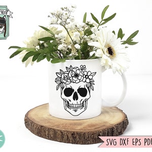 Pu&ograve; includere: Una tazza in ceramica bianca con un designo di teschio nero e una ghirlanda di fiori. La tazza &egrave; appoggiata su un disco di legno e circondata da fiori bianchi.