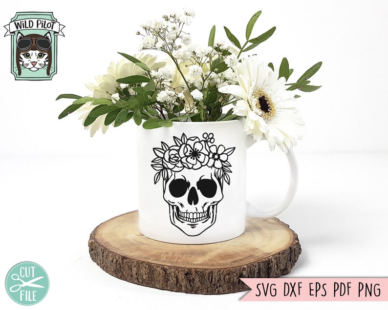 Pu&ograve; includere: Una tazza in ceramica bianca con un designo di teschio nero e una ghirlanda di fiori. La tazza &egrave; appoggiata su un disco di legno e circondata da fiori bianchi.