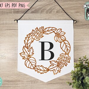 May include: A white banner with a brown glitter-like leaf wreath with the letter "B" in the center. The banner is hanging from a wooden dowel rod.