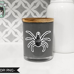 May include: A gray candle jar with a wooden lid and a black spider sticker on the front. The sticker is a simple line drawing of a spider with eight legs.
