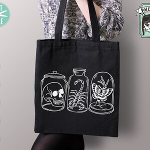 May include: A black tote bag with a white design of three glass jars containing a skull, a scorpion, and a butterfly. The text "WILD PILOT" is in a banner above the jars.