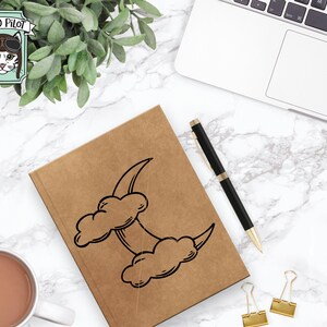 May include: A brown notebook with a black and white illustration of a crescent moon and clouds. The notebook is on a white marble surface with a pen and paper clips.
