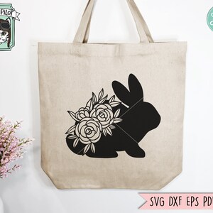 Op de afbeelding: Beige canvas draagtas met een zwart silhouet van een konijn met bloemaccenten. De tas heeft twee handvatten en de tekst "SVG DXF EPS PDF PNG" onderaan. Een logo met een kat met een pilotenbril staat in de linkerbovenhoek.