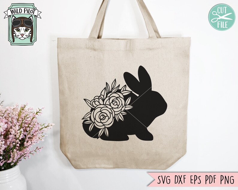 Op de afbeelding: Beige canvas draagtas met een zwart silhouet van een konijn met bloemaccenten. De tas heeft twee handvatten en de tekst "SVG DXF EPS PDF PNG" onderaan. Een logo met een kat met een pilotenbril staat in de linkerbovenhoek.