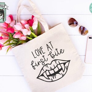 Może przedstawiać: Biała torba typu tote bag z napisem "LOVE AT first bite" i czarno-białą ilustracją ust wampira z kłami. Torba jest otoczona r&oacute;żowymi i białymi tulipanami.