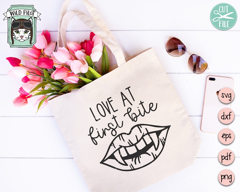 Może przedstawiać: Biała torba typu tote bag z napisem "LOVE AT first bite" i czarno-białą ilustracją ust wampira z kłami. Torba jest otoczona r&oacute;żowymi i białymi tulipanami.