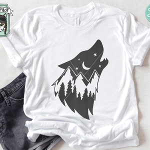 May include: A white t-shirt with a black silhouette of a wolf's head howling at the moon. The wolf's head is filled with a mountain range and pine trees. The moon is a crescent shape with stars around it.
