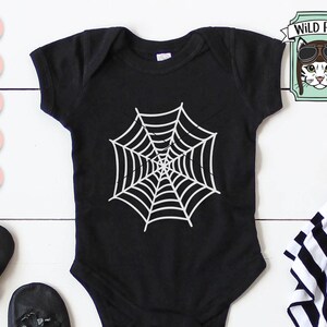 May include: Black baby onesie with a white spiderweb design. The onesie is on a white wooden surface.