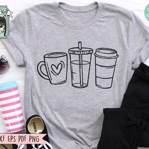 May include: A grey t-shirt featuring a design of three coffee cups. The cups are black and white and have a simple design. The design includes a mug with a heart, a tall cup with a straw, and a takeaway cup with a lid.