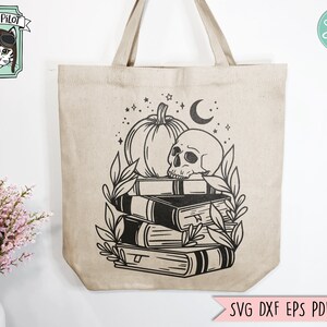 May include: Beige tote bag featuring a black graphic of a pumpkin, skull, books, and foliage. The bag has a Wild Pilot logo and the text "SVG DXF EPS PDF PNG". The bag has two handles.