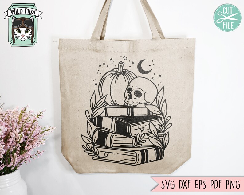 May include: Beige tote bag featuring a black graphic of a pumpkin, skull, books, and foliage. The bag has a Wild Pilot logo and the text "SVG DXF EPS PDF PNG". The bag has two handles.