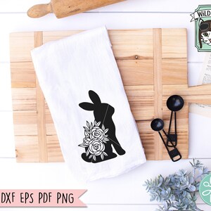 Op de afbeelding: Witte keukendoek met een zwart silhouet van een konijn en bloemenontwerp. De doek ligt op een houten snijplank met maatlepels en een deegroller. De tekst "SVG DXF EPS PDF PNG" staat onderaan.