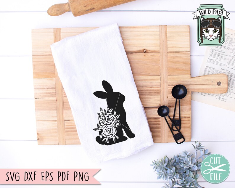Op de afbeelding: Witte keukendoek met een zwart silhouet van een konijn en bloemenontwerp. De doek ligt op een houten snijplank met maatlepels en een deegroller. De tekst "SVG DXF EPS PDF PNG" staat onderaan.