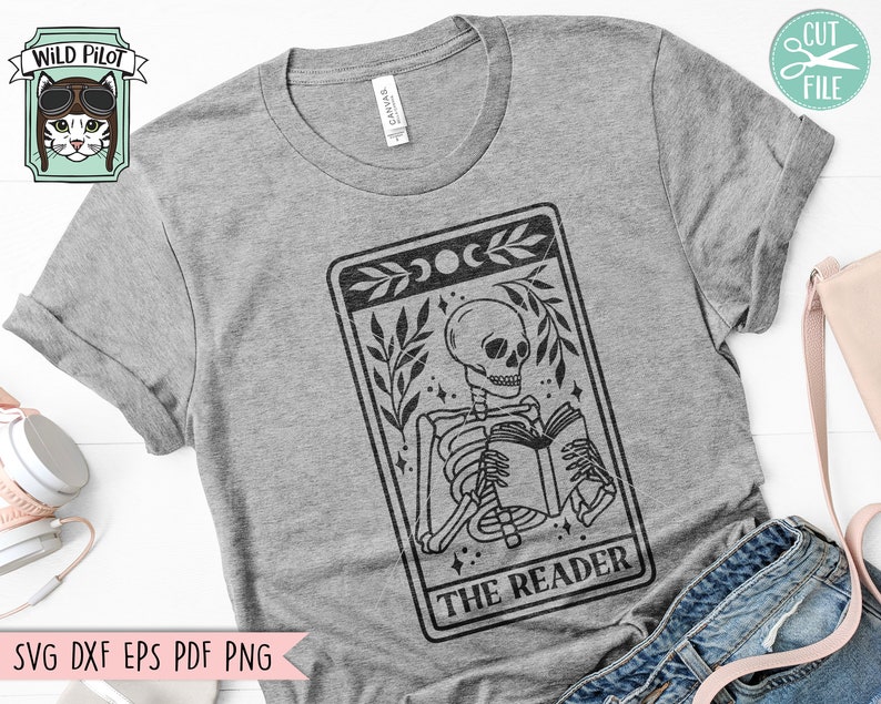 May include: A grey t-shirt with a black and white graphic of a skeleton reading a book, labelled "THE READER." The design includes crescent moons and botanical elements. The Wild Pilot logo is in the upper left corner.