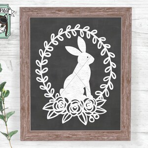 May include: A white bunny rabbit silhouette inside a wreath of leaves with white flowers. The design is on a black chalkboard background inside a brown wooden frame.