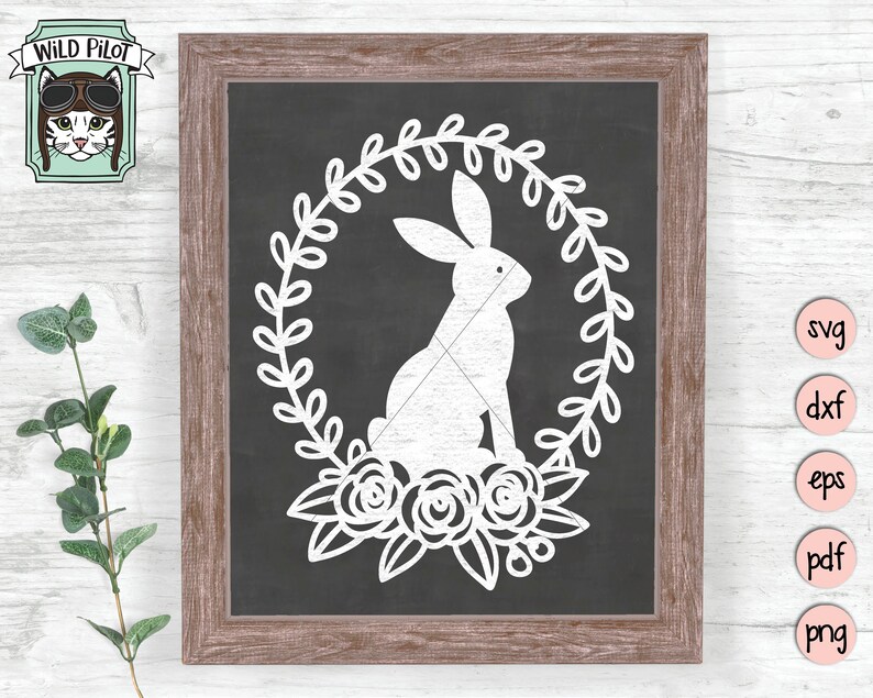 May include: A white bunny rabbit silhouette inside a wreath of leaves with white flowers. The design is on a black chalkboard background inside a brown wooden frame.