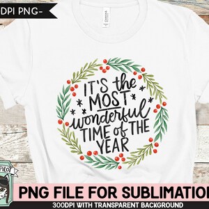 May include: White t-shirt featuring a watercolor wreath with green leaves and red berries. The text "IT'S the MOST wonderful TIME of THE YEAR" is in black script. A Wild Pilot logo is in the lower left corner.