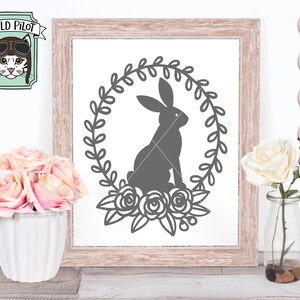 May include: A gray bunny rabbit silhouette inside a wreath of leaves with three roses at the bottom. The image is in a wooden frame with a white background.