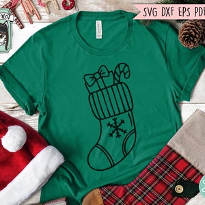 Op de afbeelding: Een groen T-shirt met een zwart-witte illustratie van een kerstsok met een strik, een snoepstok en een sneeuwvlok.