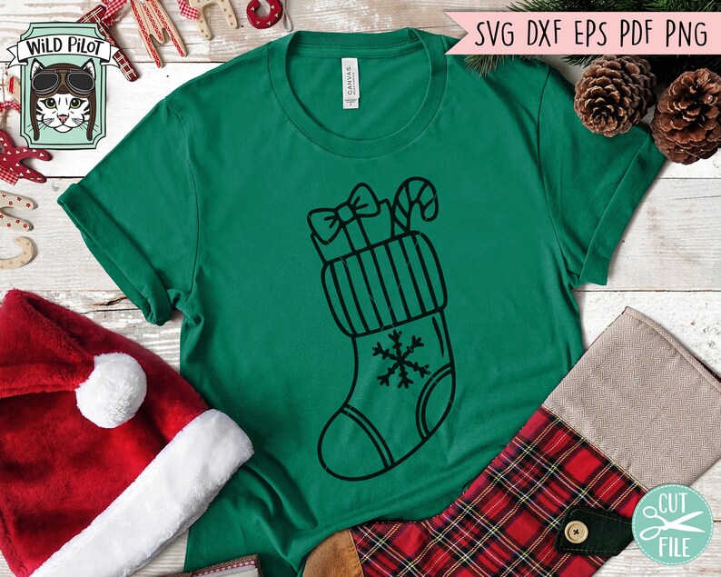 Op de afbeelding: Een groen T-shirt met een zwart-witte illustratie van een kerstsok met een strik, een snoepstok en een sneeuwvlok.