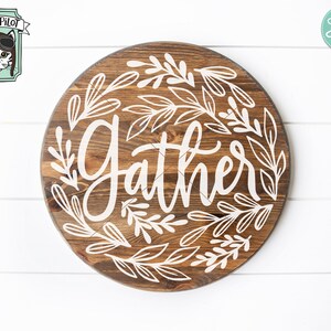 Op de afbeelding: Een rond houten bord met het woord "Gather" in witte cursieve letters omringd door witte gestileerde bladeren.