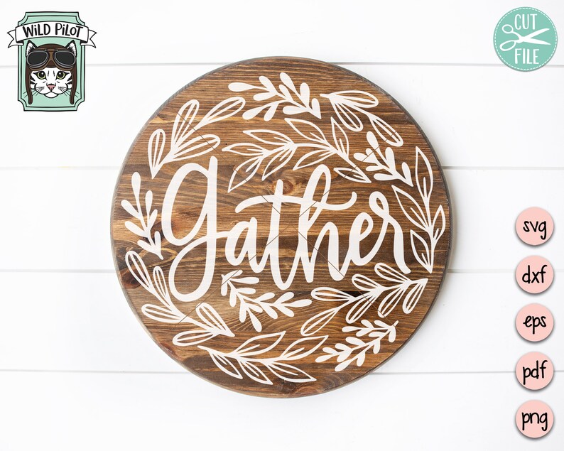 Op de afbeelding: Een rond houten bord met het woord "Gather" in witte cursieve letters omringd door witte gestileerde bladeren.