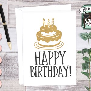 Puede incluir: Una tarjeta blanca con el texto "Happy Birthday!" en letras negras. Un pastel dorado con tres velas encendidas est&aacute; encima del texto.