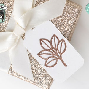 May include: A gold glitter gift box with a white satin ribbon and a white tag with a brown leaf design. The tag also has the text "CUT FILE" in a pink circle with scissors.