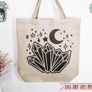 May include: A beige tote bag with a black design of crystals, a crescent moon, and stars. The design is centered on the bag.