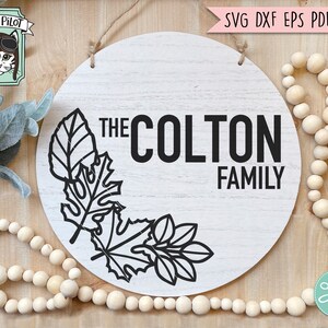 May include: A white round wooden sign with the text "The Colton Family" in black lettering. The sign is decorated with a black outline of leaves in the bottom left corner.