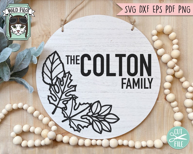 May include: A white round wooden sign with the text "The Colton Family" in black lettering. The sign is decorated with a black outline of leaves in the bottom left corner.