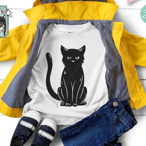 May include: A white t-shirt with a black silhouette of a cat sitting with its tail curled around its legs. The t-shirt is folded and laid on top of a yellow jacket with black stripes. A pair of blue jeans and a pair of blue sneakers are also in the image.