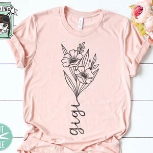 May include: A pink t-shirt with a black line drawing of a bouquet of flowers and the word "grow" written vertically. The shirt is on a white wooden surface.