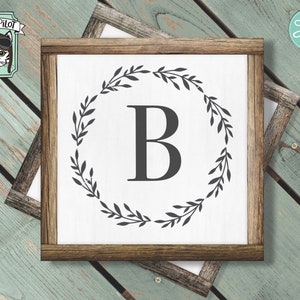 May include: A white sign with a brown wooden frame, featuring a black wreath with leaves and the letter "B" in the center.